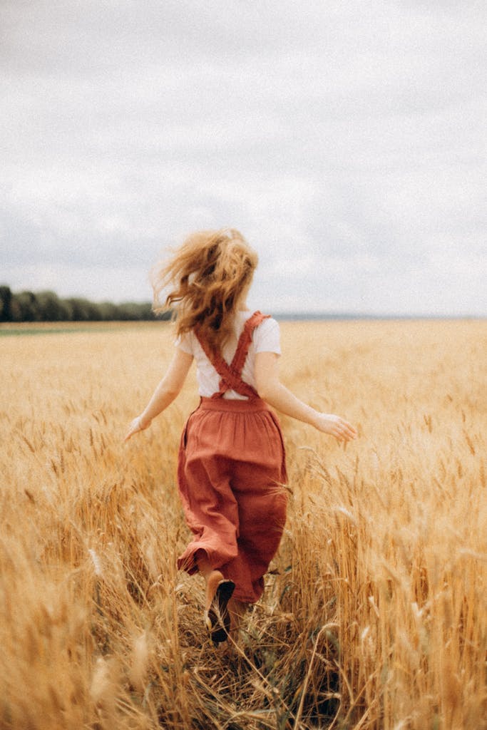 A carefree woman in a red skirt runs through a golden wheat field under a cloudy sky.