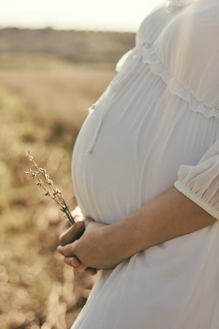 Expectant mother in white dress holds wildflowers, symbolizing new beginnings.