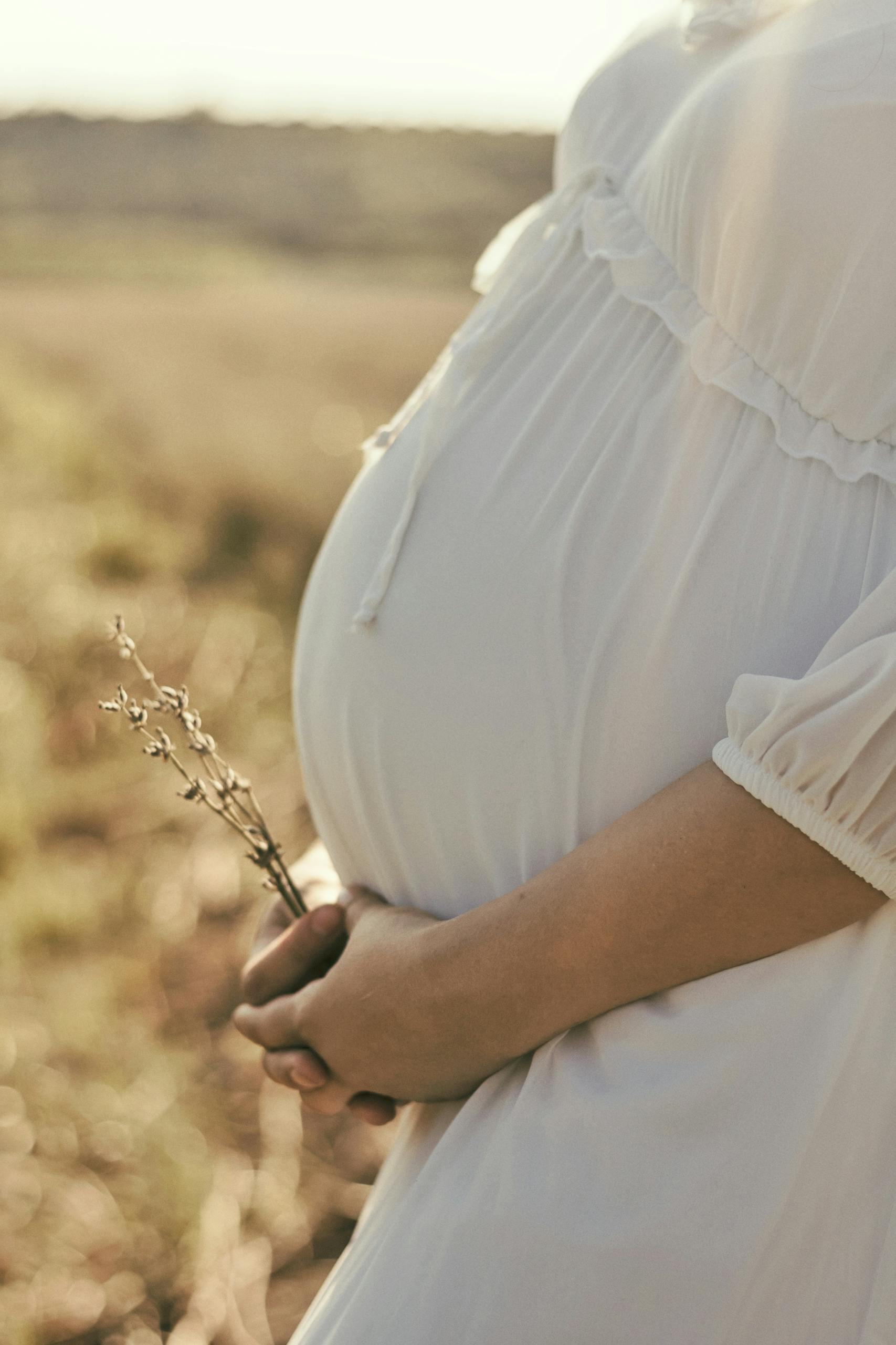 Expectant mother in white dress holds wildflowers, symbolizing new beginnings.